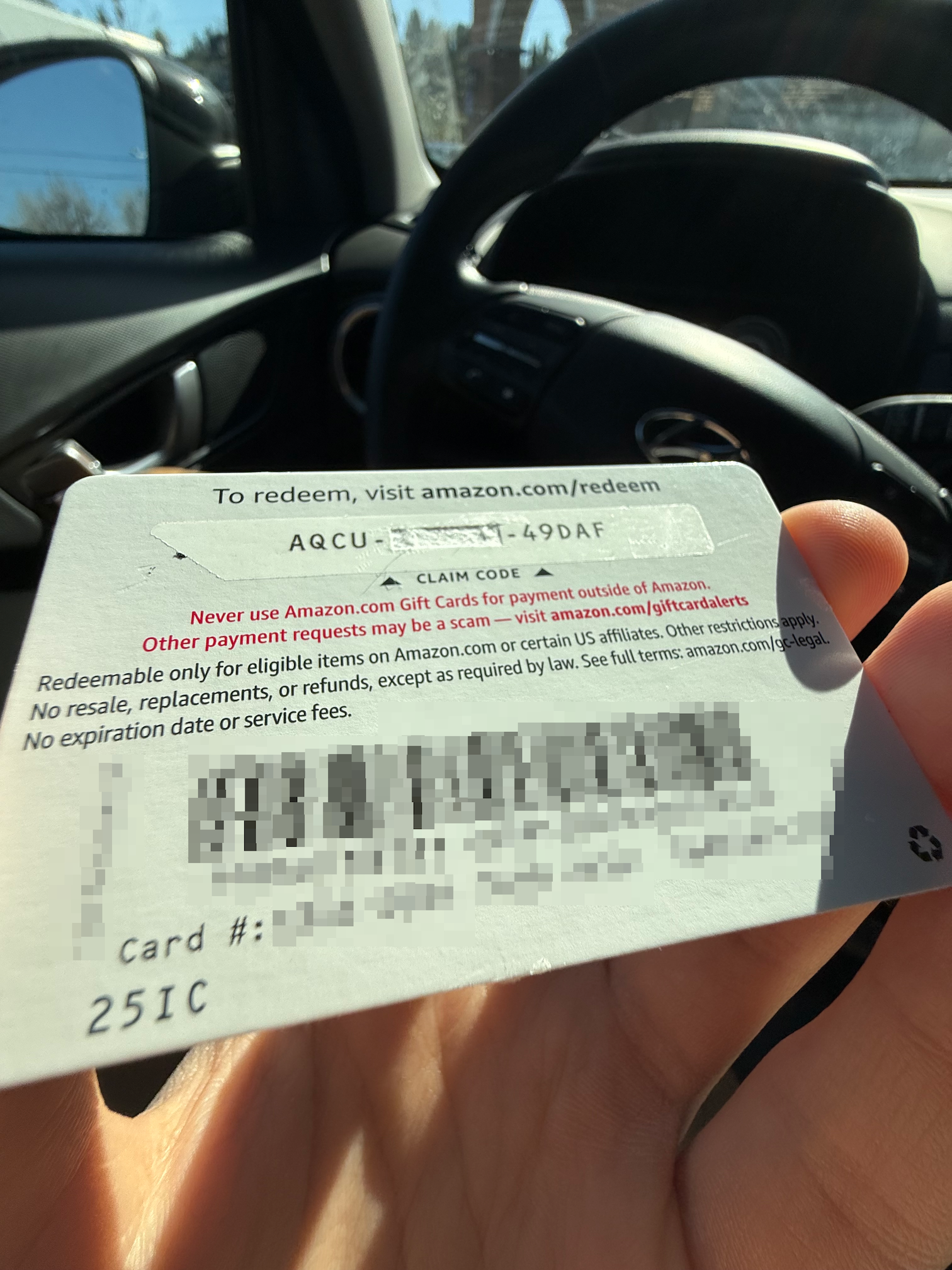 photo taken in car, hand holding amazon gift card. Claim code starts with AQCU, ends with 49DAF, middle section removed. Card number and barcode blurred out.